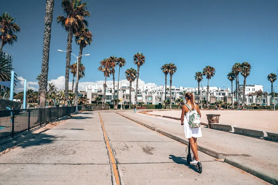 Woman enjoying a bleisure trip, riding an electric scooter along a palm-lined promenade on a sunny day.