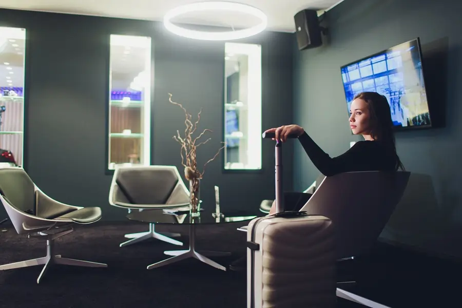Woman sitting in a modern airport lounge with a suitcase, watching a wall-mounted TV.